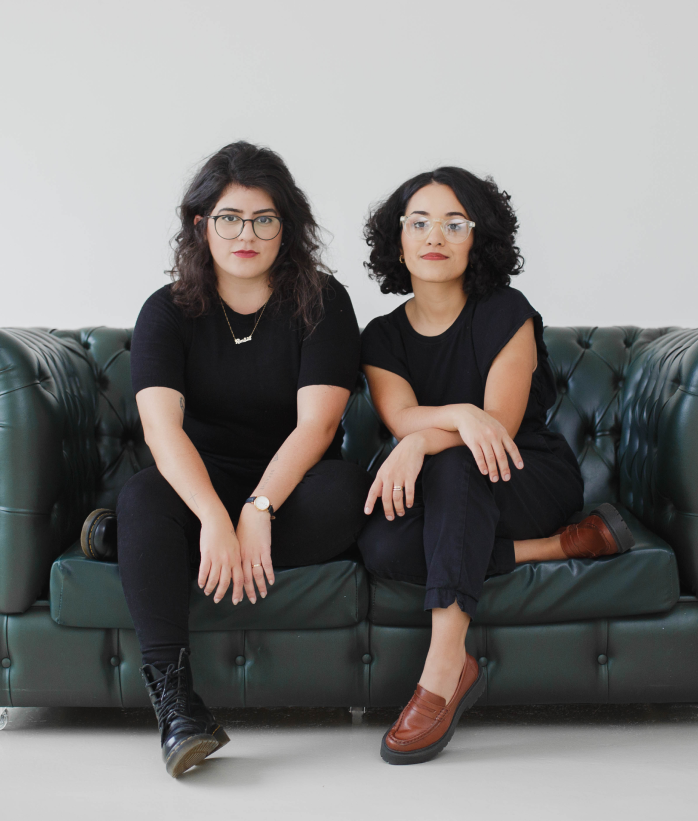 Foto da Marina e Duda sentadas em um sofá vestidas de preto.